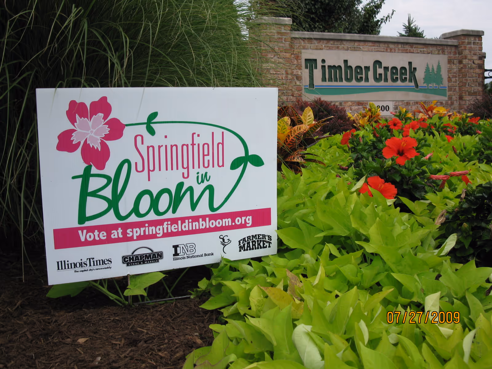 A garden area with lush green plants and red flowers in front of a brick sign that reads 'Timber Creek'. In the foreground, there is a white sign with a pink flower graphic that says 'Springfield in Bloom' and encourages voting at springfieldinbloom.org. The date 07/27/2009 is visible in the bottom right corner.