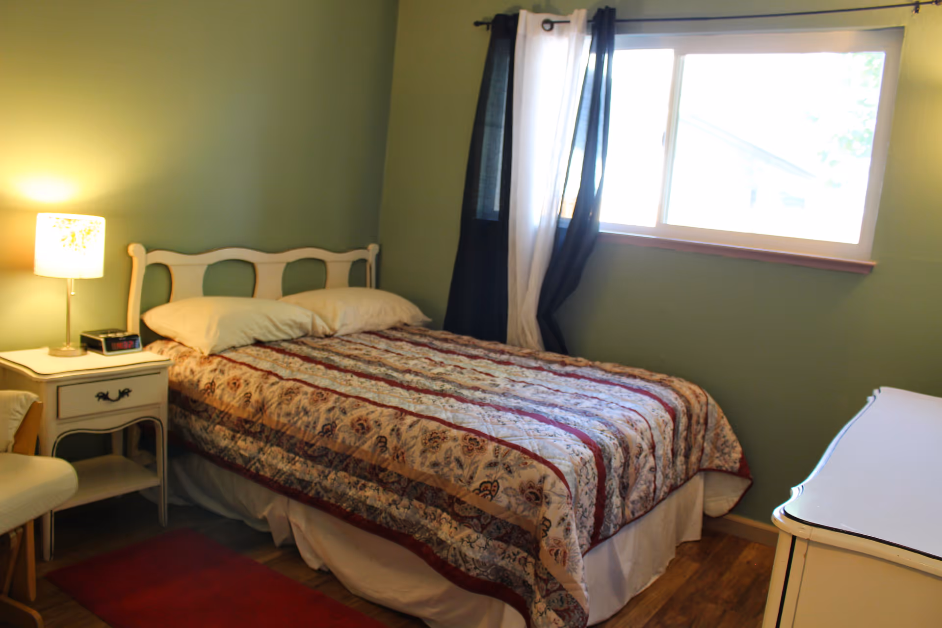 A cozy bedroom with a double bed covered in a patterned quilt. There are two pillows on the bed, a white nightstand with a lamp and an alarm clock, a white chair, and a window with black and white curtains letting in natural light. The walls are painted green and the floor is wooden.