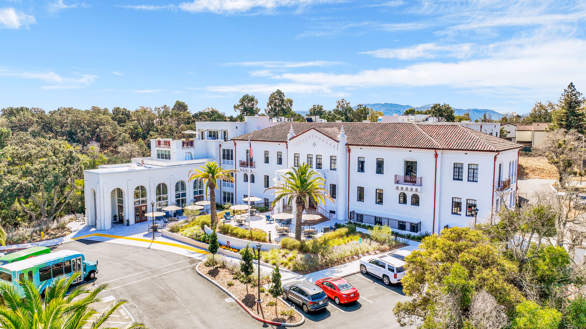 A large white multi-story senior living facility building with red roof tiles and red trim, surrounded by trees and landscaping. There are outdoor seating areas with tables and umbrellas near the entrance, several parked cars, and a green shuttle bus in the parking lot. The sky is clear with some clouds and mountains are visible in the background.