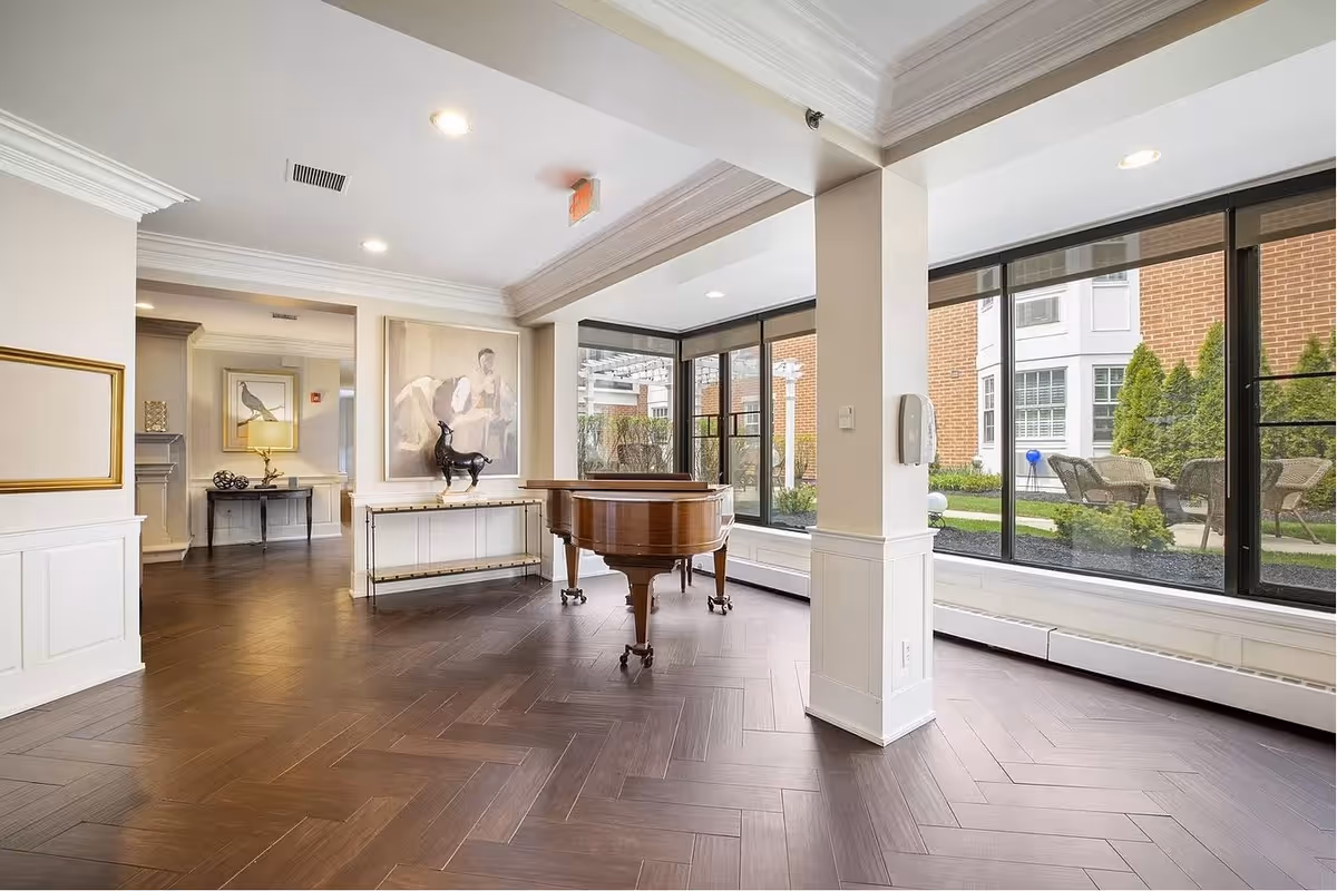 Bright and spacious interior room with large windows overlooking a garden area. The room features a wooden grand piano on a dark herringbone-patterned floor, white walls with wainscoting, and decorative artwork on the walls. Outside, there are patio chairs and greenery visible through the windows.