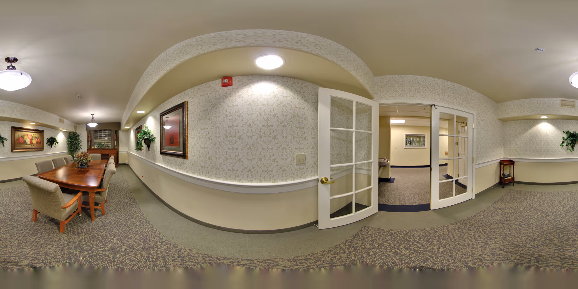 A well-lit interior hallway and dining area in a senior living facility. The dining area features a wooden table with six upholstered chairs, decorative plants, and framed artwork on the walls. The hallway has patterned wallpaper, carpeted floors, and double glass doors leading to another room.