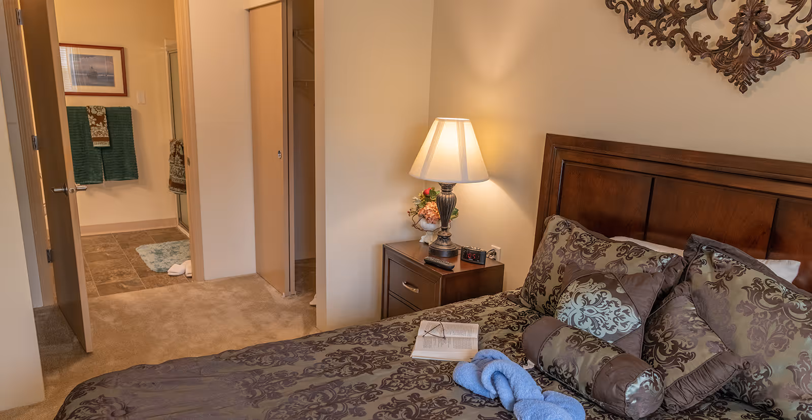 A cozy bedroom with a wooden headboard, decorative pillows, and a patterned bedspread. A nightstand with a lamp, a clock, a remote control, and a small flower arrangement is beside the bed. An open door reveals a bathroom with green towels hanging on the wall and a tiled floor.