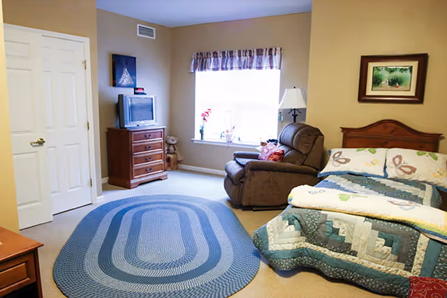 A cozy bedroom with a bed covered in a patchwork quilt, a brown recliner chair next to a window with a floral valance, a wooden dresser with a small TV on top, a floor lamp, and a framed picture on the wall.