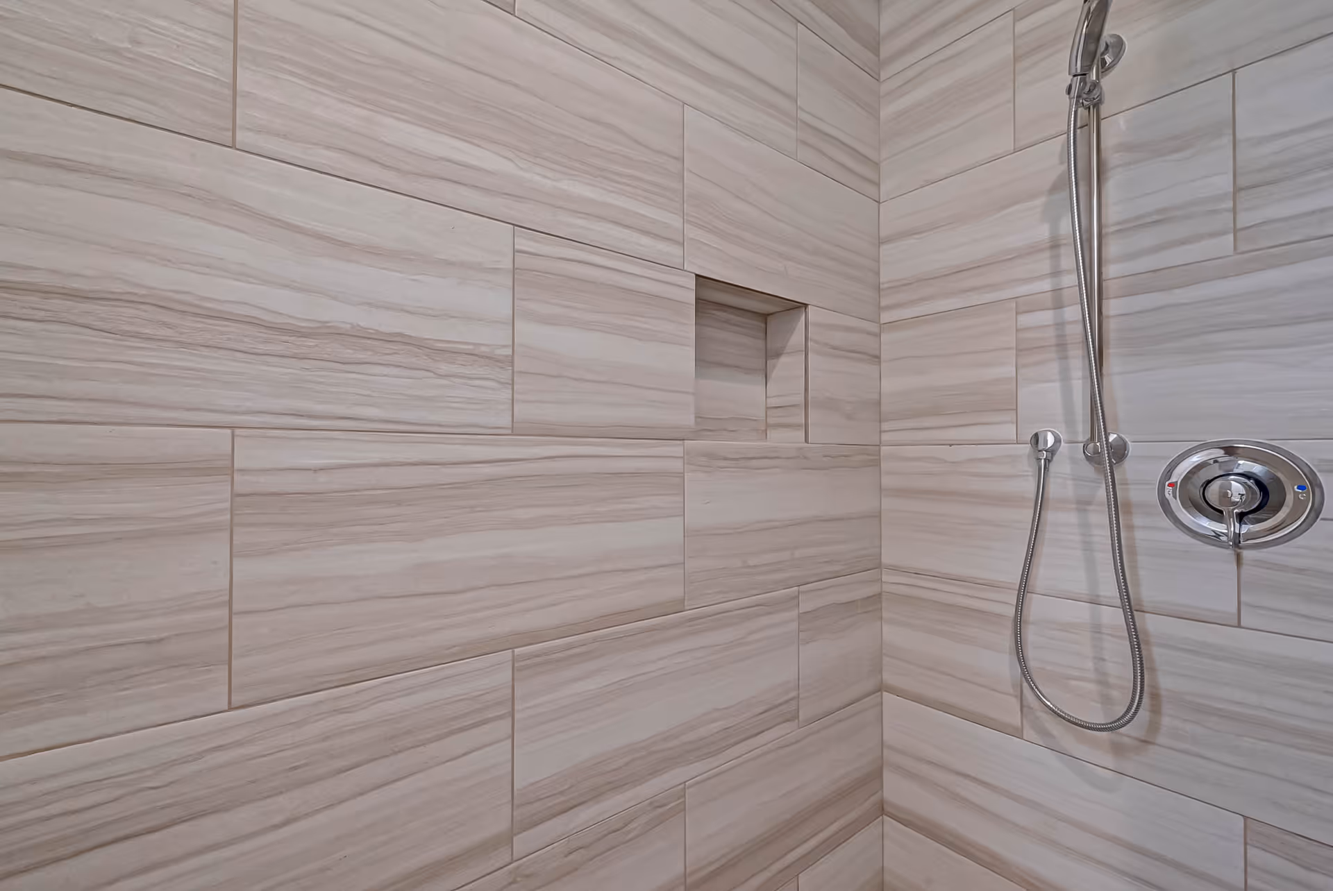 Tiled shower interior with a recessed shelf and a chrome handheld showerhead and control.