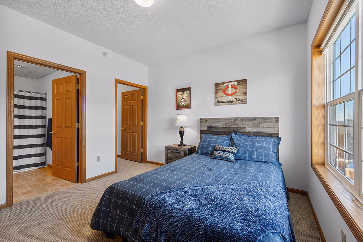 Sunlit bedroom with a blue-dressed bed, wooden headboard and nightstand with a lamp, a large window, and an open doorway showing a bathroom with a striped shower curtain.