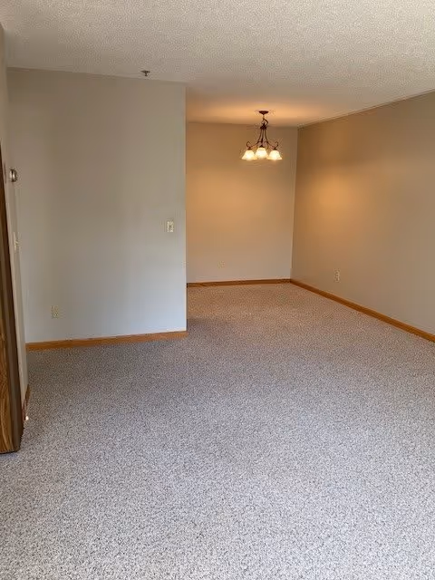Empty carpeted interior room with beige walls and a hanging dining light fixture in the far corner.