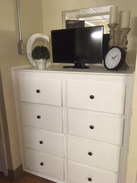 White eight-drawer dresser topped with a small TV, clock, candles, and a potted plant in an interior room.