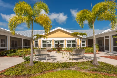 Sunny courtyard with palm trees, benches, a central fountain, and a yellow single-story building.
