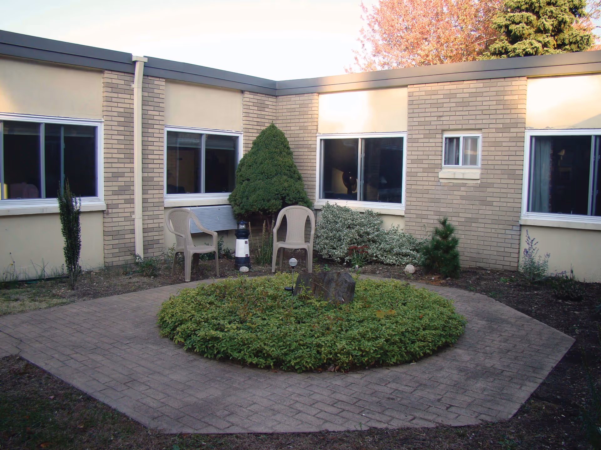 A small outdoor courtyard area with a circular garden bed in the center surrounded by a paved walkway. Two plastic chairs are placed near a trimmed bush against a beige brick building with several windows.