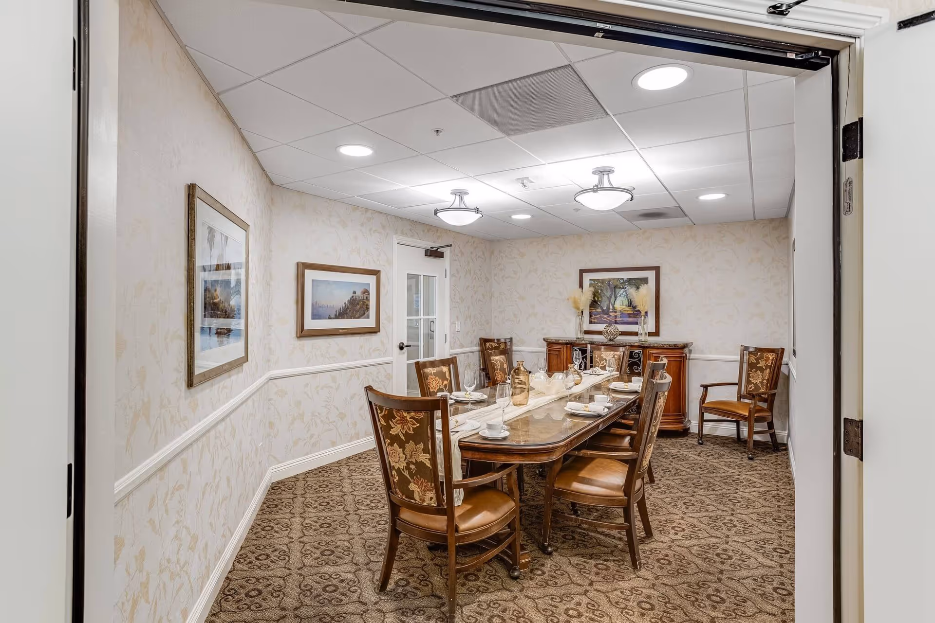 A formal dining room with a long wooden table set for six people with plates, cups, glasses, and decorative items. The room has patterned carpet, wallpaper with a subtle floral design, framed artwork on the walls, and a wooden sideboard with decorative vases and a framed picture above it.