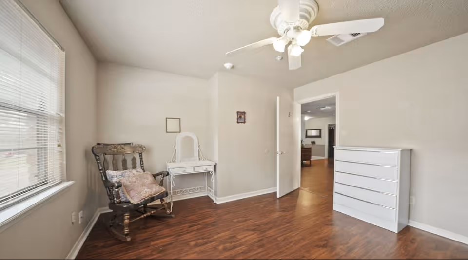 A simple bedroom with wooden flooring, a white ceiling fan with lights, a wooden rocking chair with cushions near a window with blinds, a small white vanity table with a mirror against the wall, and a white chest of drawers. The door is open, showing a glimpse of another room with wooden flooring.