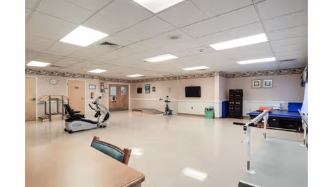 Large rehabilitation therapy room with exercise equipment, parallel bars, tables, chairs, and a wall-mounted TV.