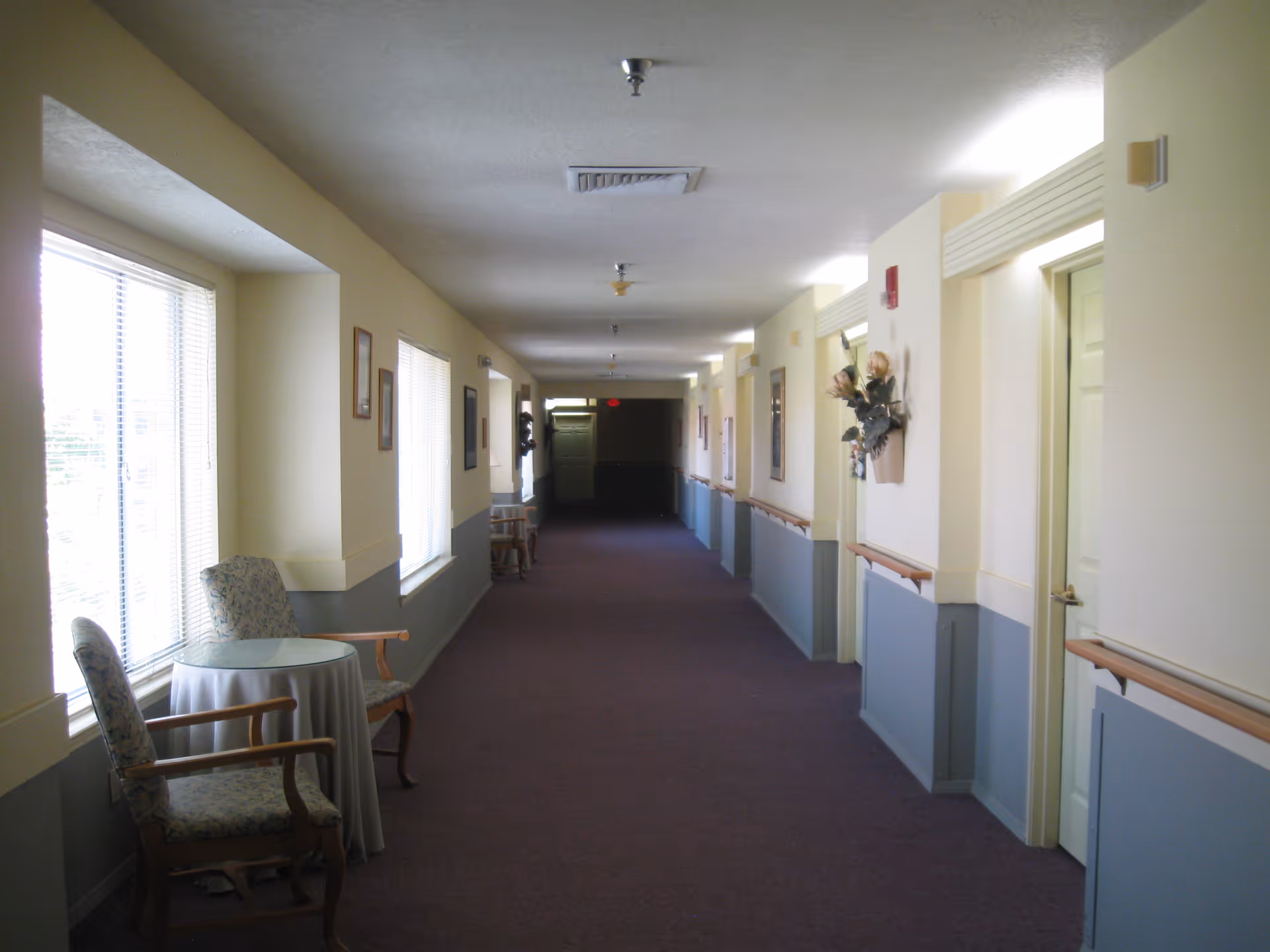 A long, well-lit hallway in an assisted living facility with beige and blue walls, carpeted floor, handrails along the walls, several closed doors on the right, and windows with blinds on the left. There are a few chairs and small tables placed near the windows, and decorative wall hangings are visible.