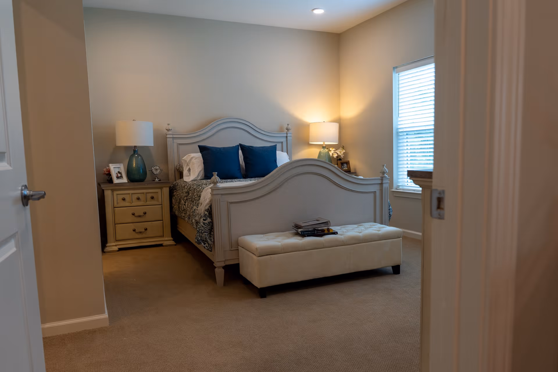 View into a bedroom with a white wooden bed frame, blue pillows, and a beige bench at the foot of the bed. There are two nightstands with lamps on either side of the bed, a window with blinds on the right wall, and beige carpet flooring.