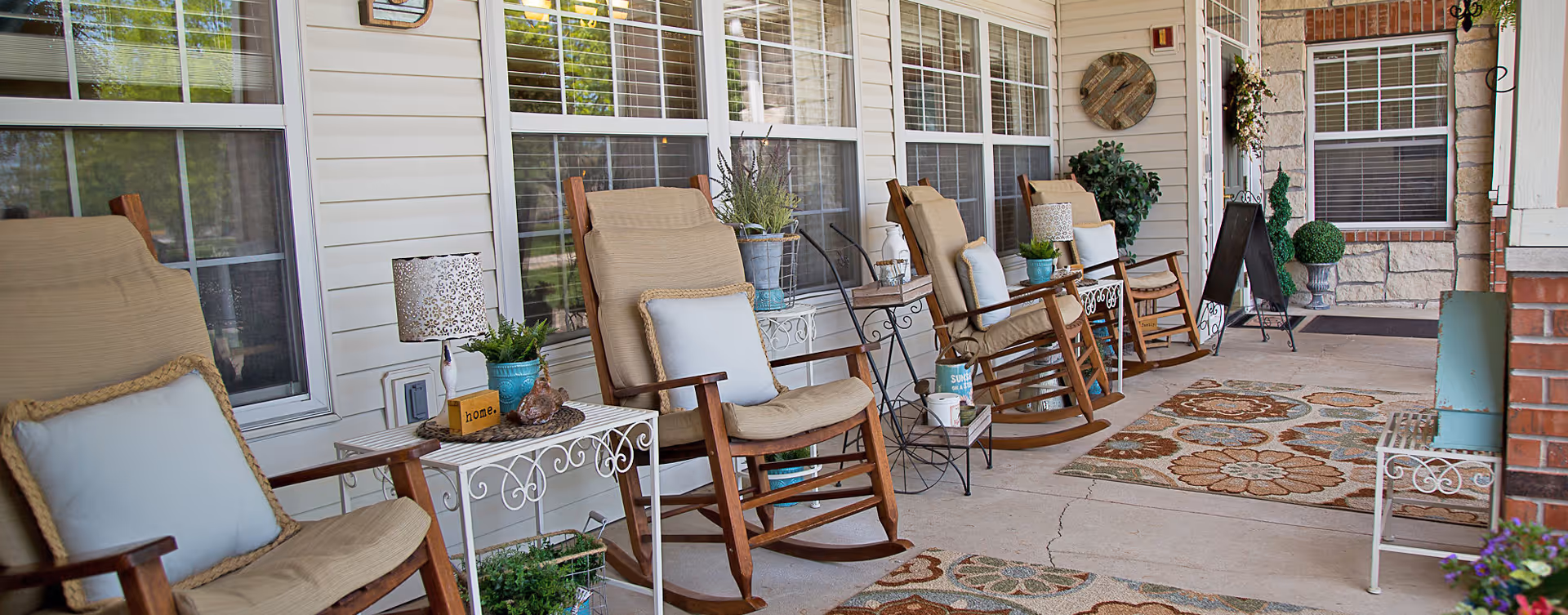 Covered front porch showing a row of cushioned wooden rocking chairs, small side tables, potted plants, and decorative rugs.