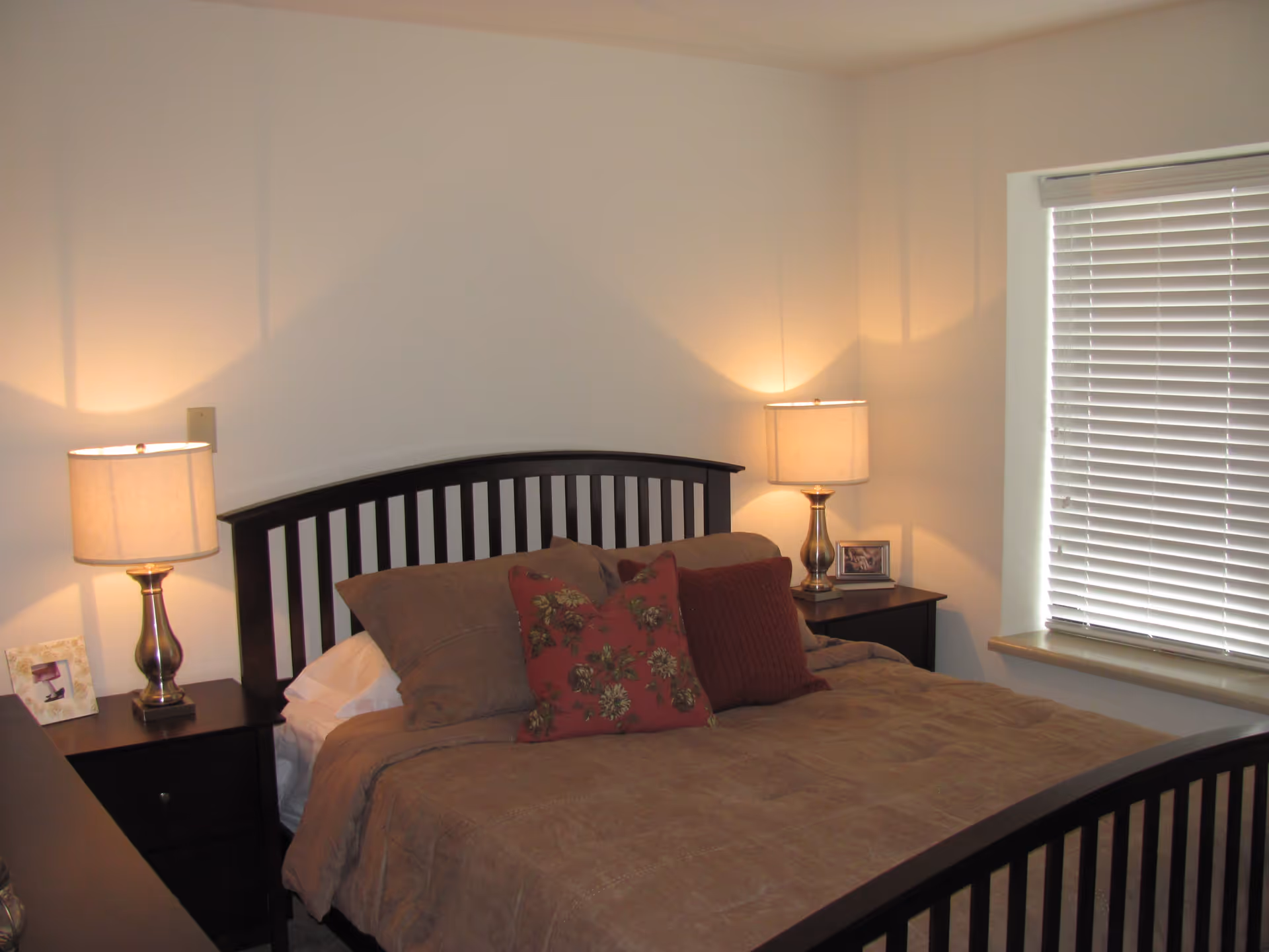 A bedroom with a dark wooden bed frame and a brown comforter. There are two bedside tables, each with a lamp and a framed photo. A window with closed blinds is on the right side of the room.