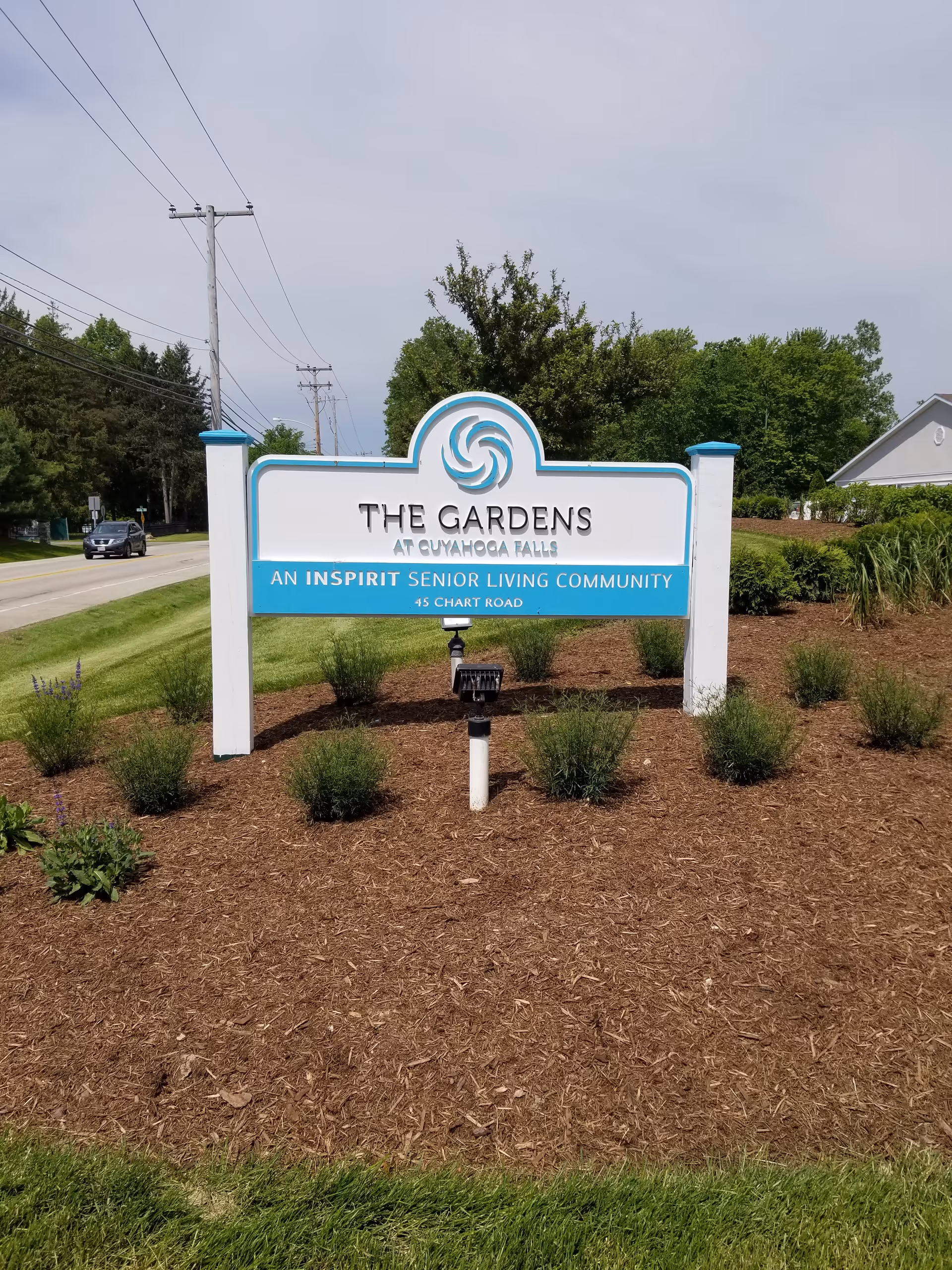 A white and blue sign for The Gardens at Cuyahoga Falls, an Inspir Senior Living Community, located at 45 Chart Road. The sign is surrounded by mulch and small bushes, with a road and trees in the background under a cloudy sky.