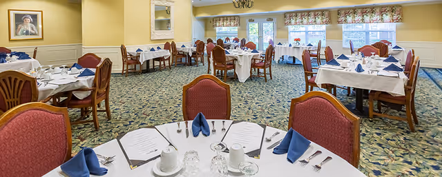 A dining room with multiple round tables covered with white tablecloths, each set with blue folded napkins, menus, cups, glasses, and silverware. The room has patterned carpet, yellow walls, floral valances on the windows, and framed artwork on the walls.