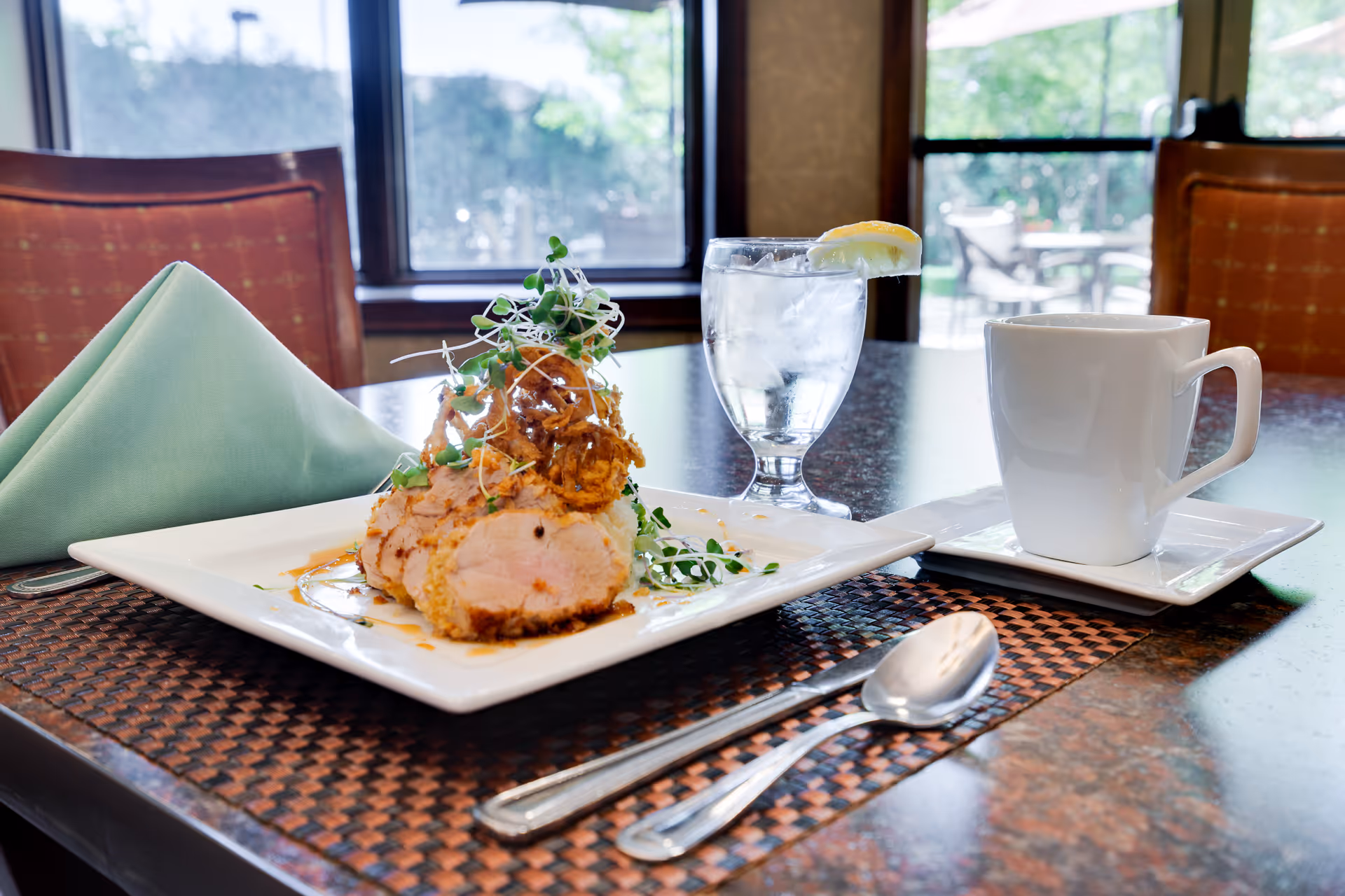 A plated meal consisting of sliced meat garnished with greens and crispy fried onions on a white square plate, placed on a woven placemat on a dining table. Next to the plate is a folded green cloth napkin, a glass of water with a lemon wedge, and a white coffee cup on a saucer. The background shows windows with a view of an outdoor patio area.