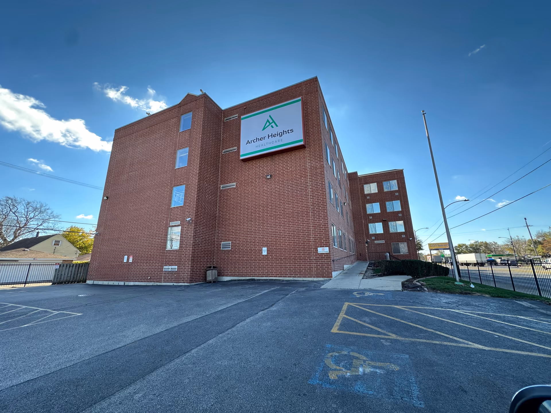 Exterior view of a multi-story brick building with a sign that reads 'Archer Heights Healthcare' on the side. The building is adjacent to a parking lot with marked handicap parking spaces and a flagpole. The sky is clear with a few clouds.