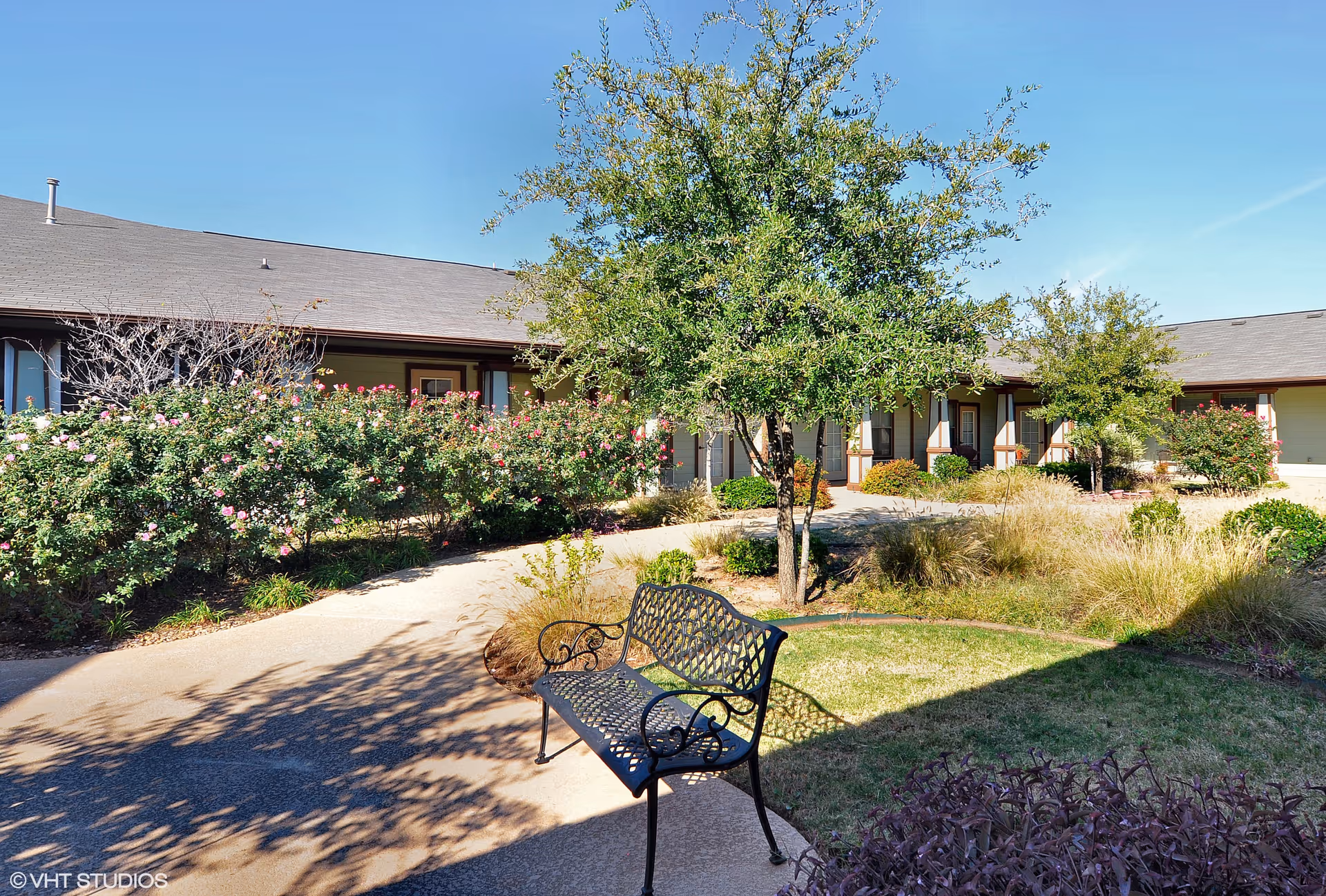 Outdoor courtyard area of Pecan Point Assisted Living and Memory Care with a metal bench on a paved walkway, surrounded by green grass, trees, and flowering bushes under a clear blue sky.