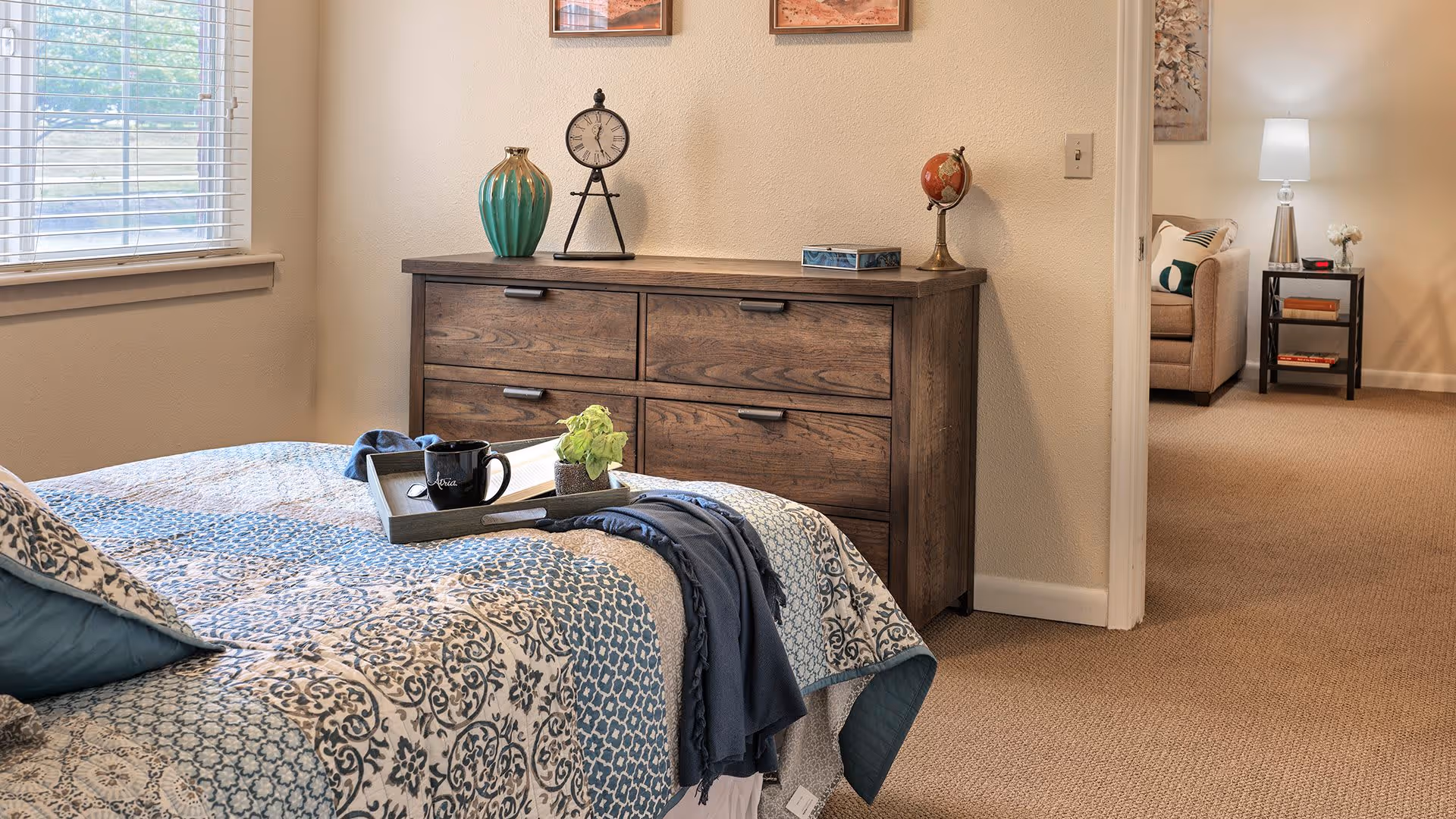 A cozy bedroom with a patterned blue and beige bedspread, a wooden dresser with decorative items including a green vase, a clock, and a small globe. A tray with a black mug, a book, and a small plant is placed on the bed. A window with blinds is on the left, and an open doorway leads to a living room area with a beige armchair, a side table with books, a lamp, and a floral wall art.