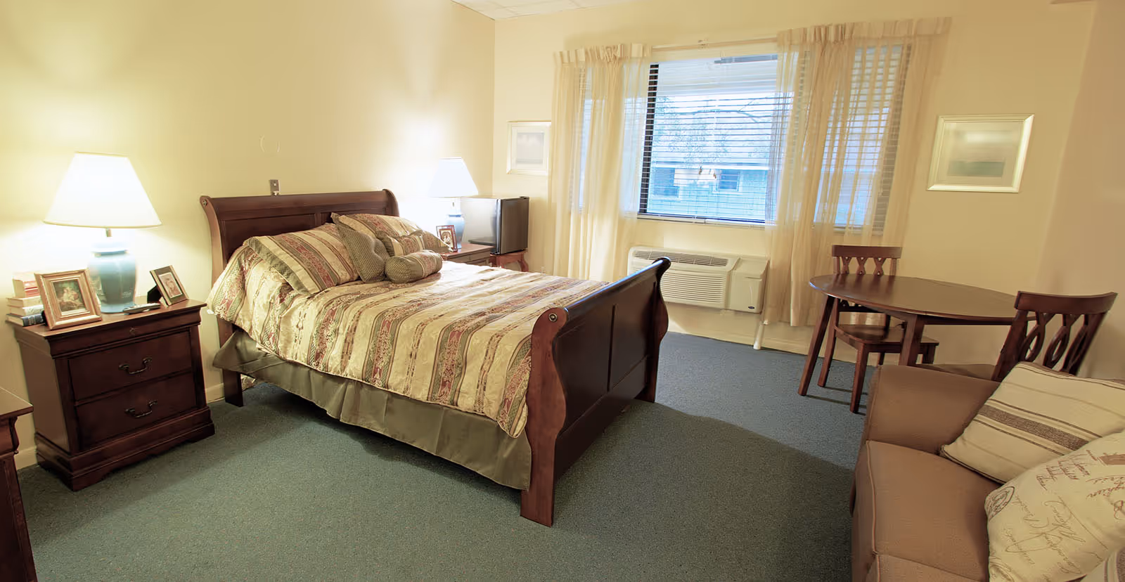 A cozy bedroom in an assisted living facility featuring a wooden bed with patterned bedding and multiple pillows. There are two bedside tables with lamps and framed photos. A window with sheer curtains lets in natural light. The room also includes a small round dining table with two chairs, a beige sofa with decorative pillows, and a wall-mounted air conditioning unit below the window.