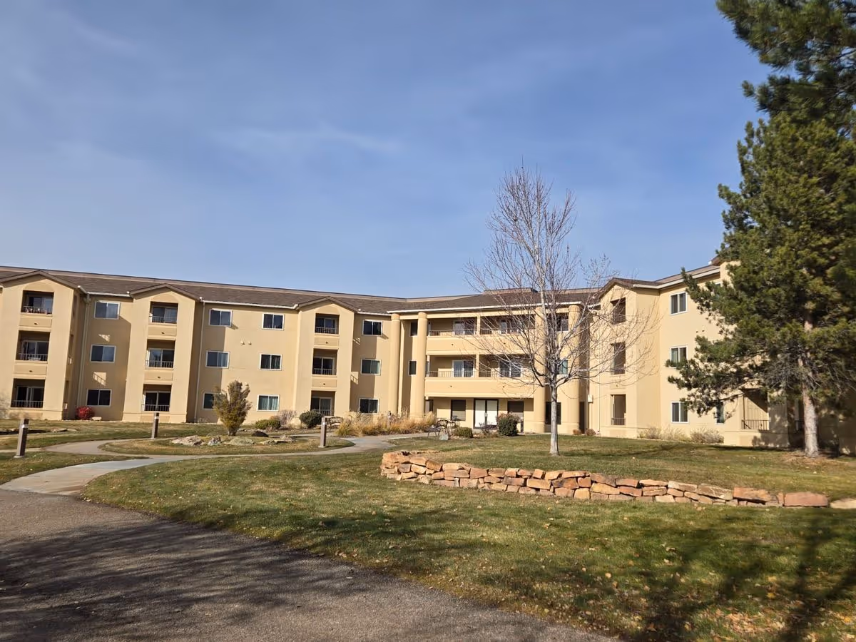 Front view of a three-story beige retirement community building with balconies, lawn, walkways, and trees.