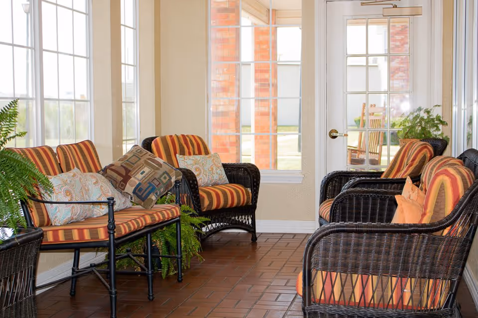 A sunlit seating area with wicker chairs and a loveseat featuring striped orange and red cushions, decorative pillows, and green potted plants. Large windows and a glass door provide a view of the outdoor area with brick pillars.