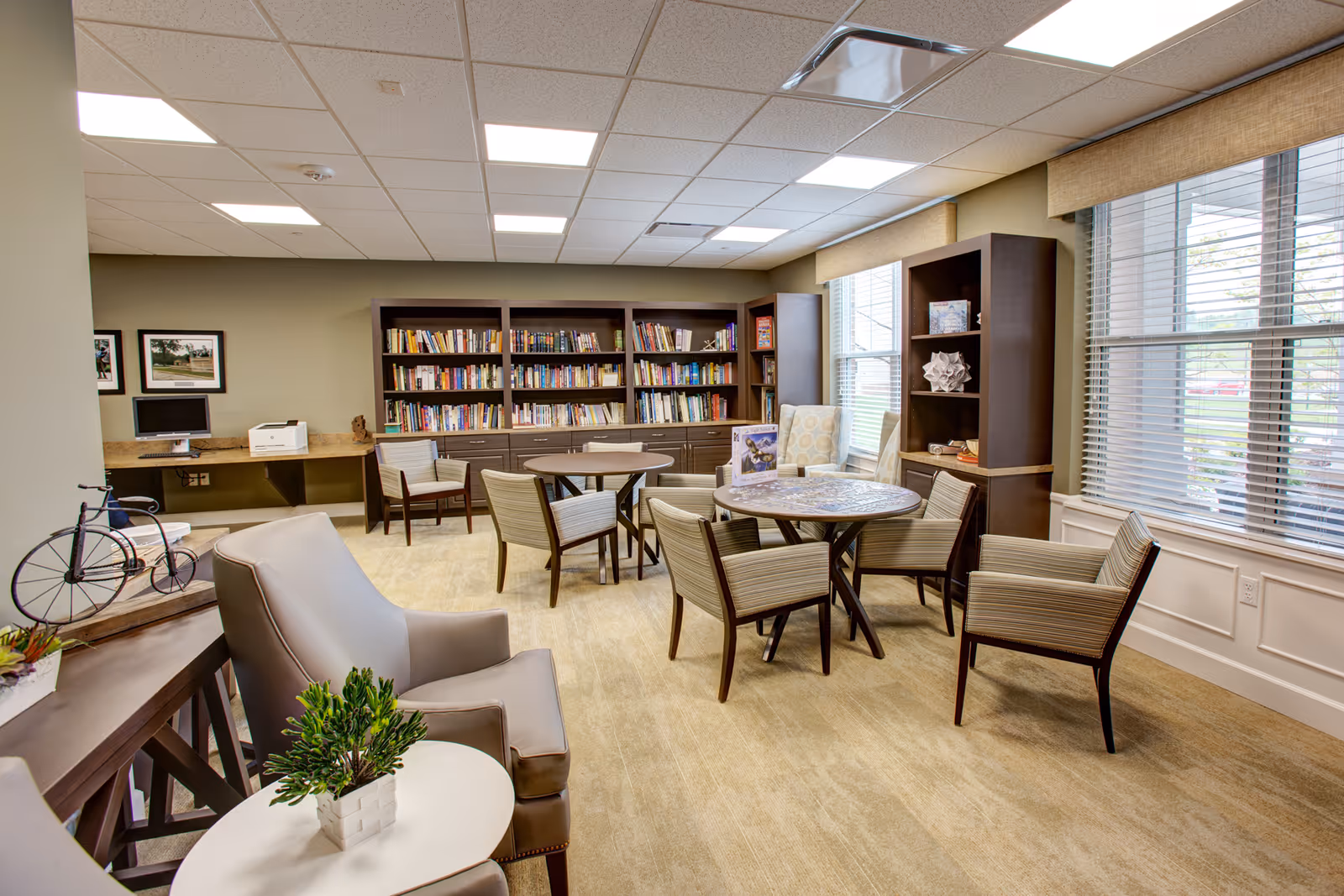 A bright and cozy common area with multiple chairs and round tables, bookshelves filled with books, large windows with blinds letting in natural light, a computer station with a printer, and decorative items including a small plant and a bicycle sculpture.