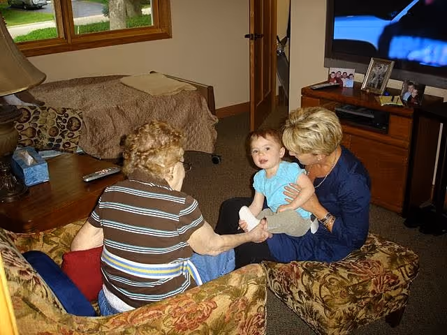 An elderly woman and a middle-aged woman sitting on floral-patterned chairs in a living room, interacting with a baby who is sitting on the middle-aged woman's lap. The room has a wooden coffee table, a couch, a television on a wooden stand, and a window showing greenery outside.