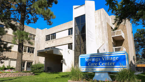Exterior view of the Springs Village Care Center building with an entrance canopy and a blue sign on a grassy lawn.