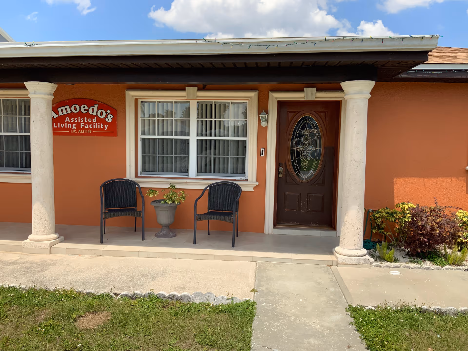 Front entrance of Amoedo's Assisted Living Facility with two white columns supporting a small roof over a porch area. There are two black chairs and a planter with greenery between them on the porch. The building exterior is painted orange with a brown door featuring decorative glass. A red sign on the wall reads 'Amoedo's Assisted Living Facility LIC. AL11149'.