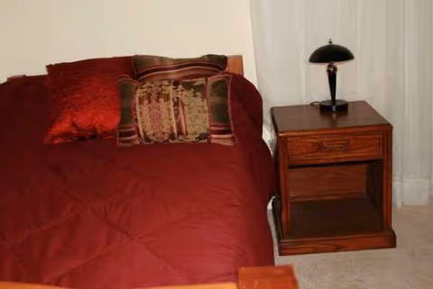 Red quilted bed with pillows and a wooden nightstand with lamp in bedroom.