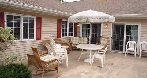 Outdoor patio area with beige siding walls, multiple windows, and sliding glass doors. The patio has a round white table with a large white umbrella, several white plastic chairs, and wicker chairs with cushions arranged around the space.