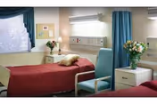 Two beds in a care facility bedroom with red comforters, nightstands, and a window.