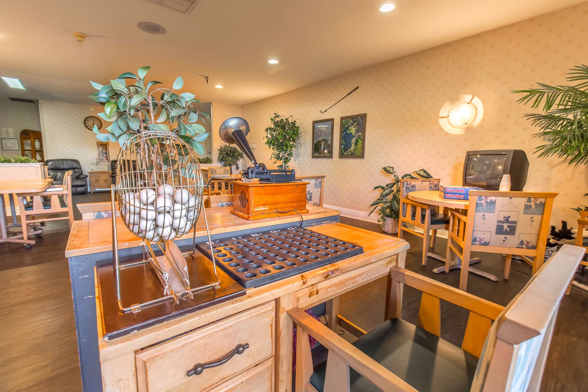 A cozy common area in Windsor Court Assisted Living featuring a bingo game setup with a cage full of bingo balls and a wooden bingo board. The room has wooden chairs and tables, a vintage gramophone, a small TV, board games, potted plants, and framed pictures on the wall, creating a warm and inviting atmosphere.