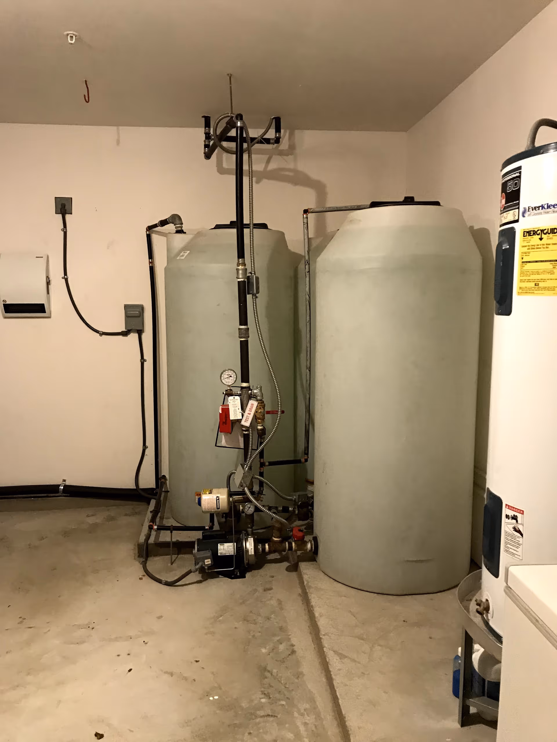 A utility room with two large cylindrical water tanks connected by pipes and a water pump on the floor. There is also a water heater partially visible on the right side of the image. The room has plain beige walls and a concrete floor.