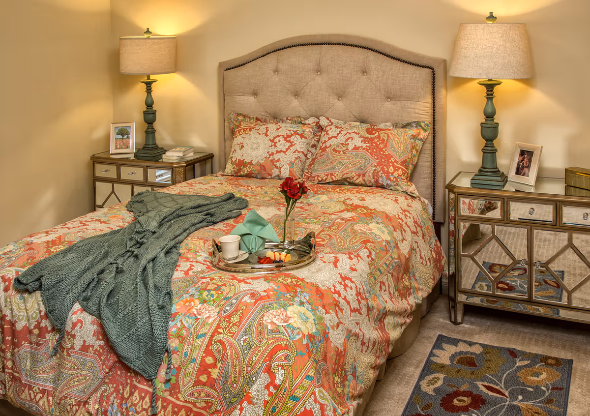 A cozy bedroom with a bed featuring a beige tufted headboard and colorful paisley-patterned bedding in shades of red, orange, and green. A green knitted throw blanket is draped over the bed. On the bed is a silver tray holding a cup, a napkin, and a small flower vase with a red flower. On either side of the bed are mirrored nightstands with green lamps, framed photos, and books. A floral-patterned rug is partially visible on the floor.