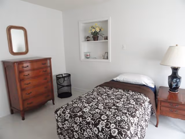 Simple bedroom with a single bed covered by a patterned blanket, a wooden dresser, nightstand with lamp, and built-in wall shelf.