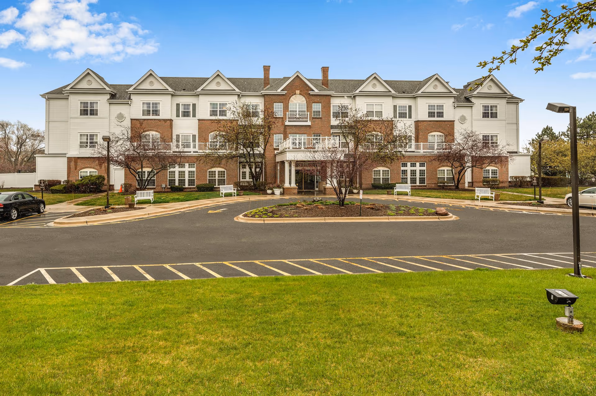 Front exterior view of a three-story senior living facility building with a combination of brick and white siding. The building has multiple windows, small balconies, and a covered entrance. There is a circular driveway with parking spaces, green grass, and a few trees in front of the building under a partly cloudy blue sky.