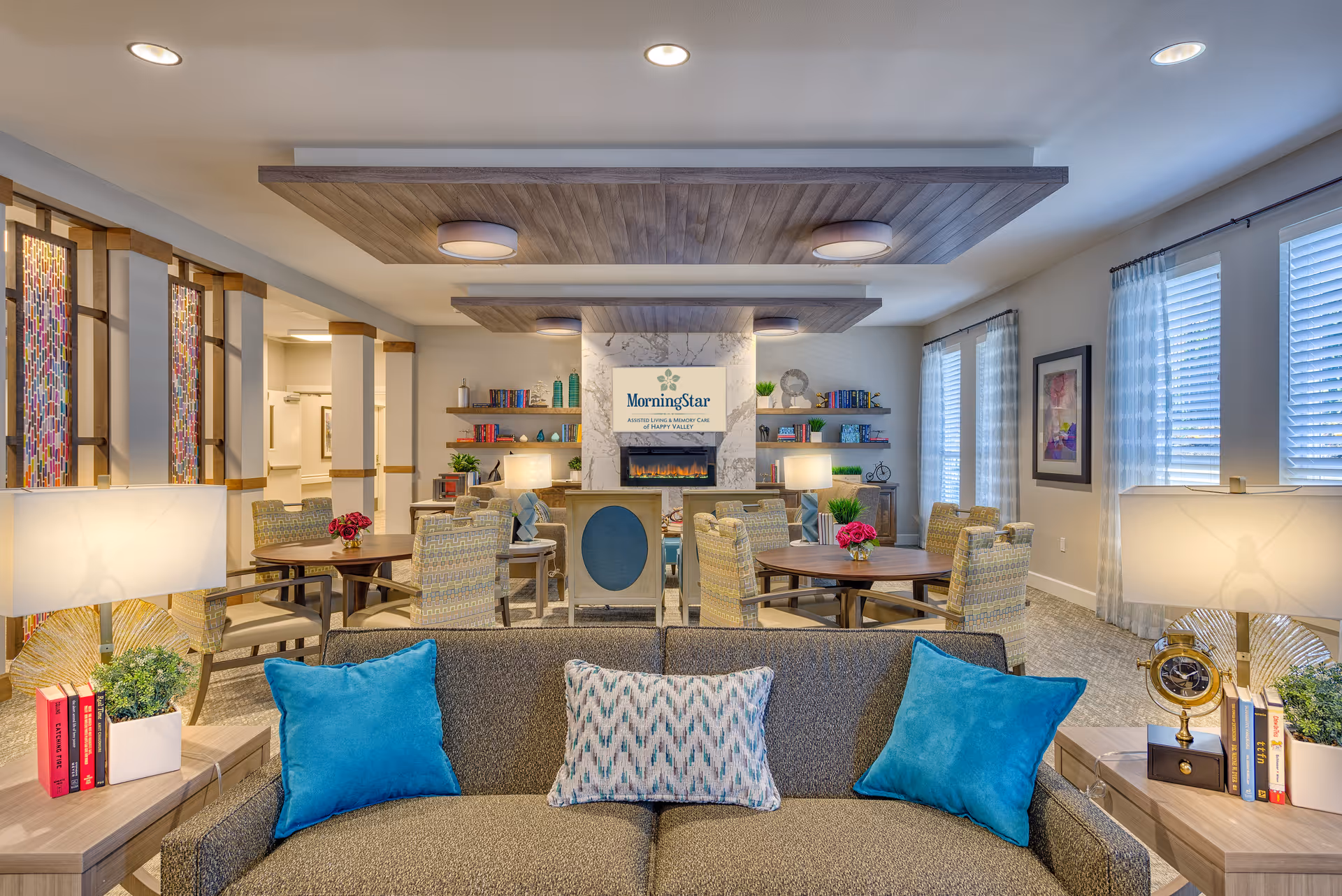 A bright and inviting common area in MorningStar Assisted Living & Memory Care of Happy Valley featuring a comfortable gray sofa with blue and patterned pillows in the foreground. Behind the sofa are round tables with cushioned chairs, decorative lamps, bookshelves with books and ornaments, and large windows with sheer curtains allowing natural light to fill the room. The ceiling has wooden panel accents with recessed lighting, and a fireplace with a MorningStar sign above it is centered on the far wall.