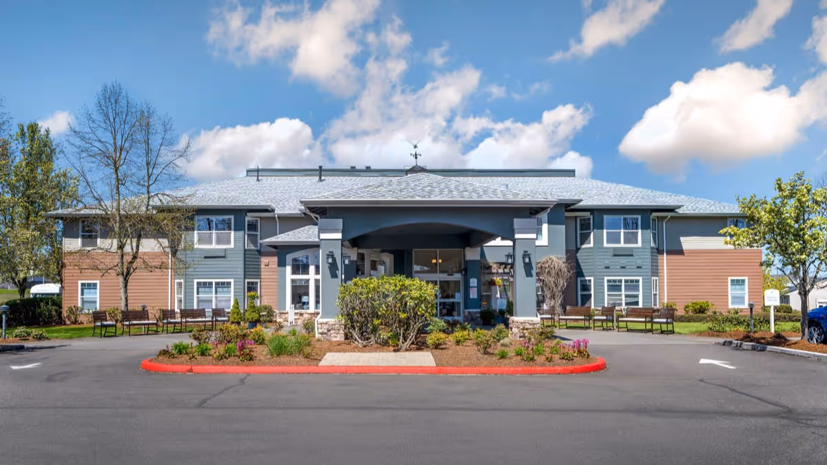 Front exterior of a two-story senior living building with a covered entrance, landscaped planter, and benches.