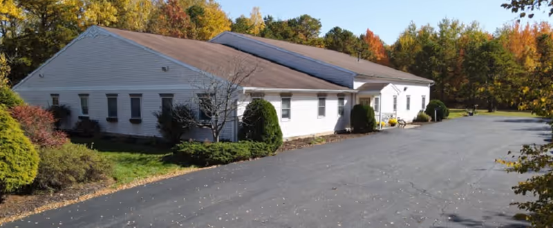 Exterior view of a single-story white building with a brown roof surrounded by trees with autumn foliage. The building is situated next to a paved driveway or parking area.