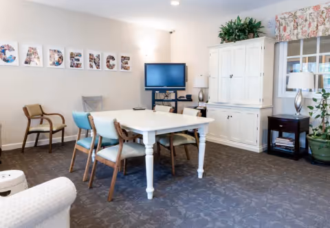 A cozy common area in a senior living facility with a white table surrounded by six chairs, a white cabinet, a TV on a stand, a side table with a lamp, and a window with floral curtains. The wall has letters spelling 'CADENCE' decorated with photos.