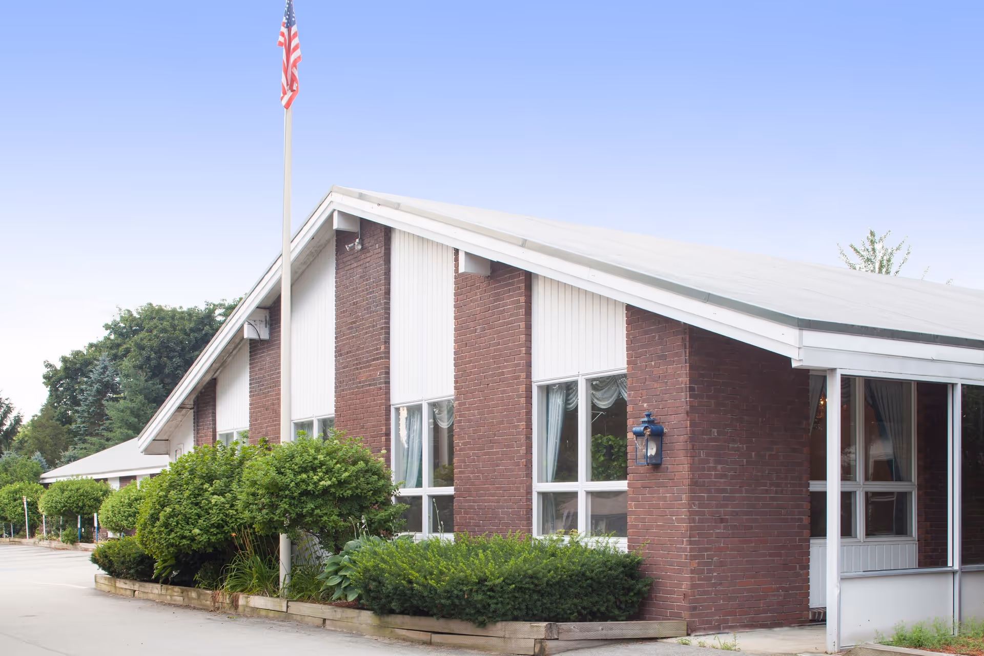 Exterior view of a single-story brick building with large windows and a sloped roof. There are green bushes and plants along the front of the building, and an American flag is mounted on a pole near the entrance. The sky is clear and blue.
