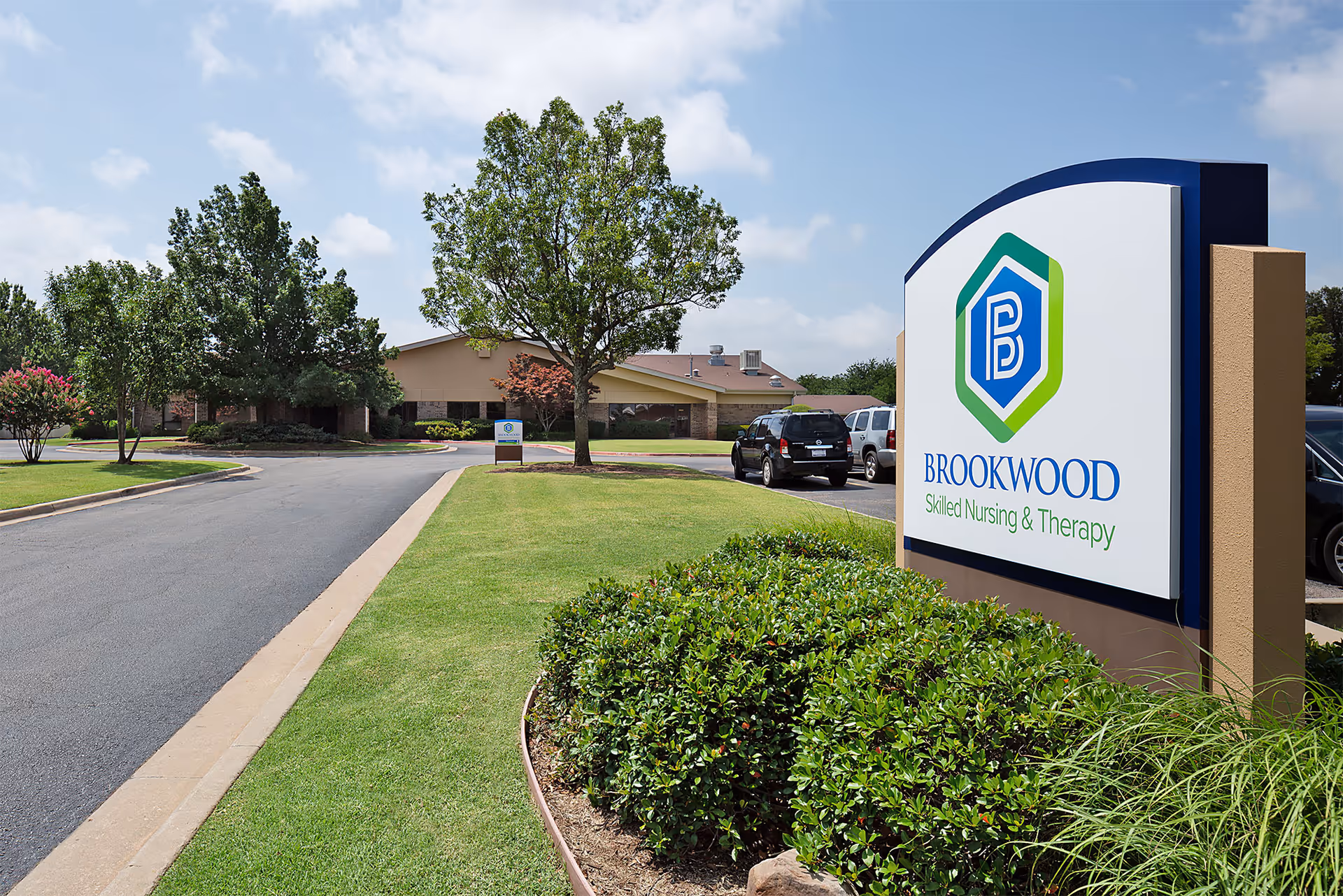 Exterior view of Brookwood Skilled Nursing & Therapy facility with a large sign in the foreground displaying the facility's name and logo. The building is surrounded by green lawns, trees, and a paved driveway with parked cars.