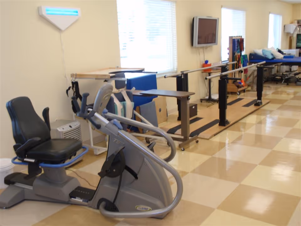 A rehabilitation room with exercise equipment including a recumbent exercise bike, parallel bars for walking practice, therapy tables, and various physical therapy tools. The room has large windows with blinds, a wall-mounted TV, and a checkered tile floor.
