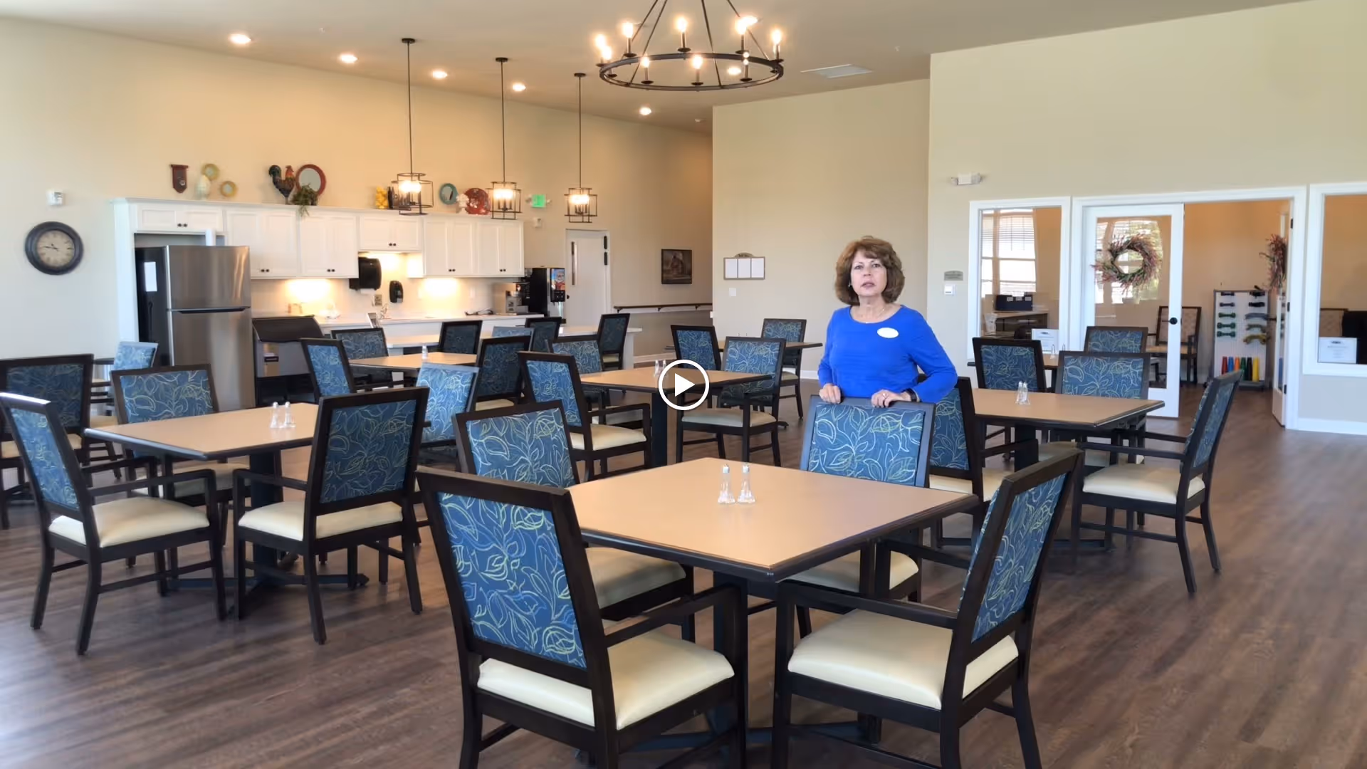 A spacious dining room in a senior living facility with multiple square tables and chairs arranged neatly. A woman in a blue top stands near one of the tables. The room features wood flooring, white cabinetry with a refrigerator and coffee station in the background, and modern light fixtures hanging from the ceiling.