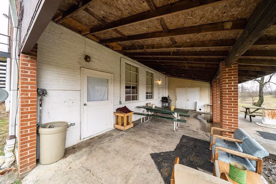 Covered outdoor patio area with a wooden ceiling and brick pillars. There is a white door and two windows on a white brick wall. The area contains a green picnic table, a grill, a trash can, and some chairs. Outside the covered area, there is a picnic table and a fire pit on a grassy field.